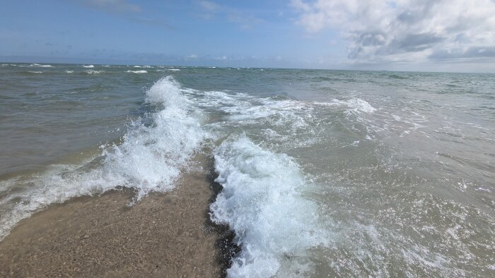 Vågor möts vid stranden i Skagen under en blå himmel med moln.