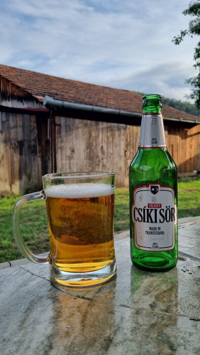 Flaska och glas med öl på bord framför träbyggnad med grönska i bakgrunden.