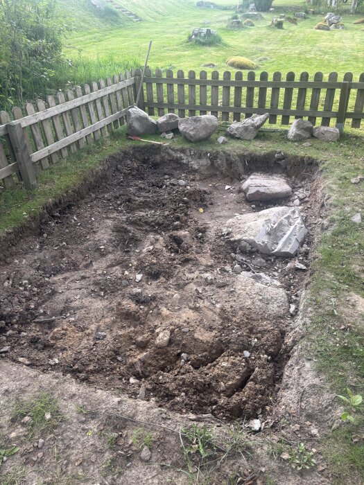 Grävd grund för växthus med synliga stenar, omgiven av trästaket och gräs.