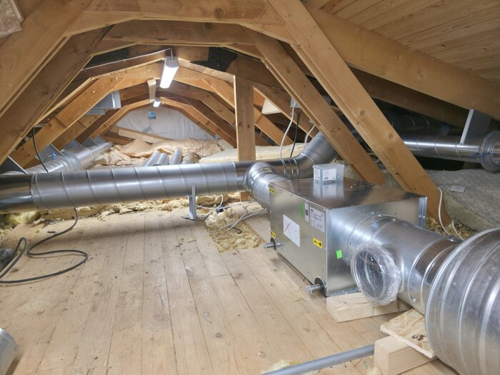 Vindsutrymme med träbjälkar och ventilationsrör synliga under taket, vilket ger inblick i byggandets konstruktion och installationer.
