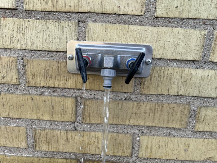 Utomhusvattenkran med läckage, monterad på gul tegelvägg, med separata vred för varm- och kallvatten.