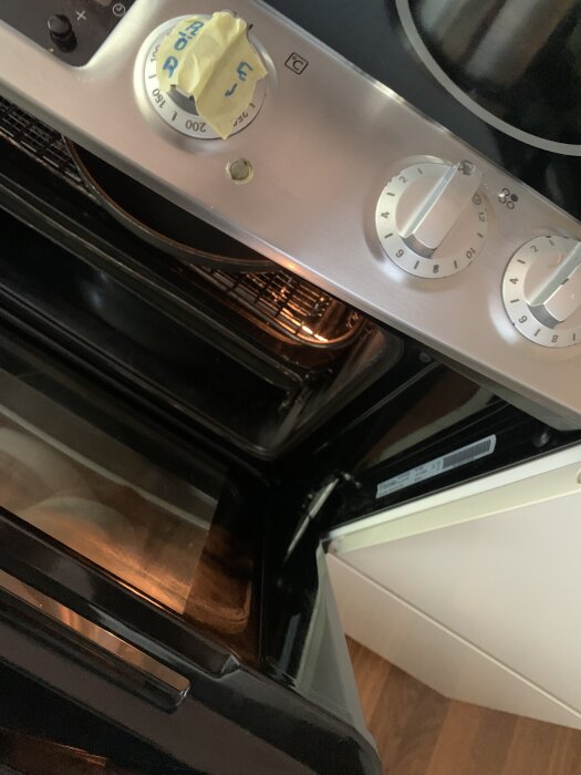Närbild av en ugn med öppen lucka och termostat, inställd på 200 grader, med ett tejp över. Lampan inuti ugnen verkar vara tänd.