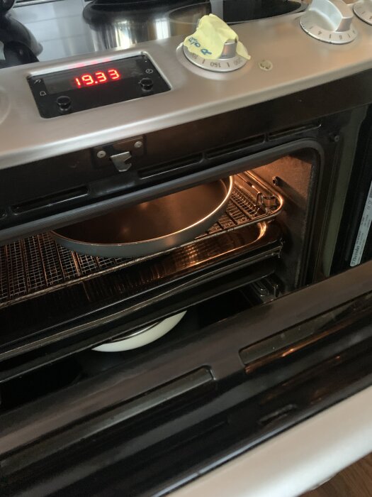 Öppen ugn med en plåt och en kastrull inuti, spisknapp markerad med tejp, termometer visar 19.33, belysning tänd.