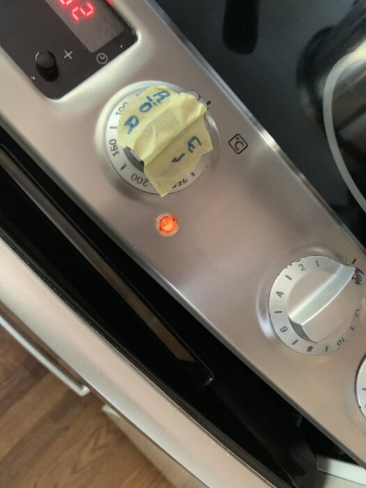 En ugns kontrollpanel med termostatvridknapp täckt av tejp och lysande indikatorlampa. Digital display visar siffror.