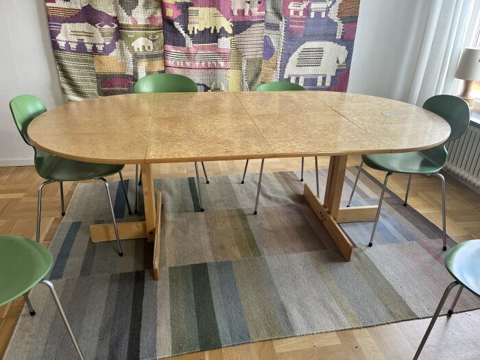 Träbord med ljus fanér, står på en randig matta omgiven av gröna stolar. Bakom syns en färgglad textilvägg med djurmotiv.
