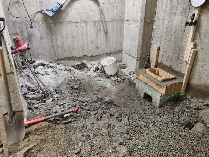 Nybetonggjutet utrymme med borttagna stenar. Verktyg och en hink syns, samt grus på marken. Brädfodring för gjutform vid väggen.