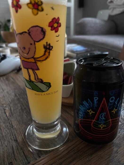 Ett glas öl bredvid en Omnipollo Omnipollos Hatt-burk på ett träbord, glaset dekorerat med en tecknad figur.