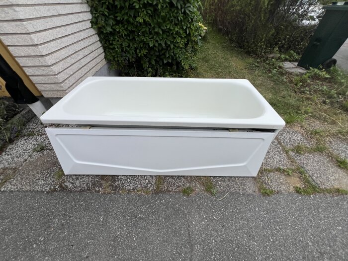 Gammalt vitt plåtbadkar med två täcksidor placerat utomhus, nära en husvägg och omgiven av grönska.