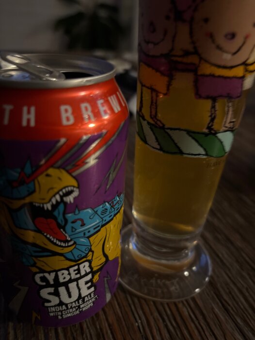 Burk med Cyber Sue IPA från Toppling Goliath Brewing och ett glas med gyllene öl, på ett rustikt träbord.