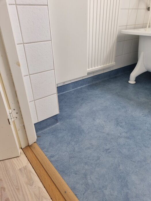 Badrumsgolv med blå matta som går upp mot kaklade väggar och vid ett hörn av ett badkar. Tröskelövergång till trä.