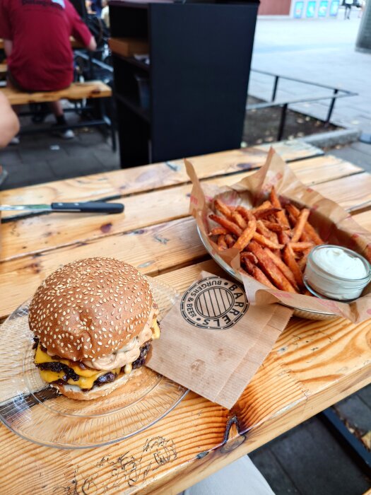 Burger with cheese and sauce on a glass plate next to a basket of sweet potato fries and dip on a wooden table at Apex x Barrels.