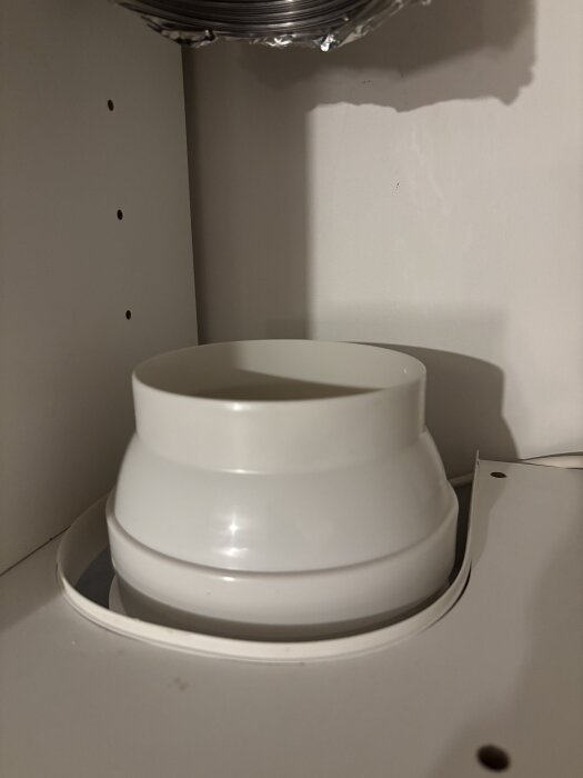 Vit plastadapter för köksfläkt, saknar gummipackning, på en 150 mm utgång. Adaptern minskar rördiametern till 115 mm, ses ovanifrån i ett skåp.