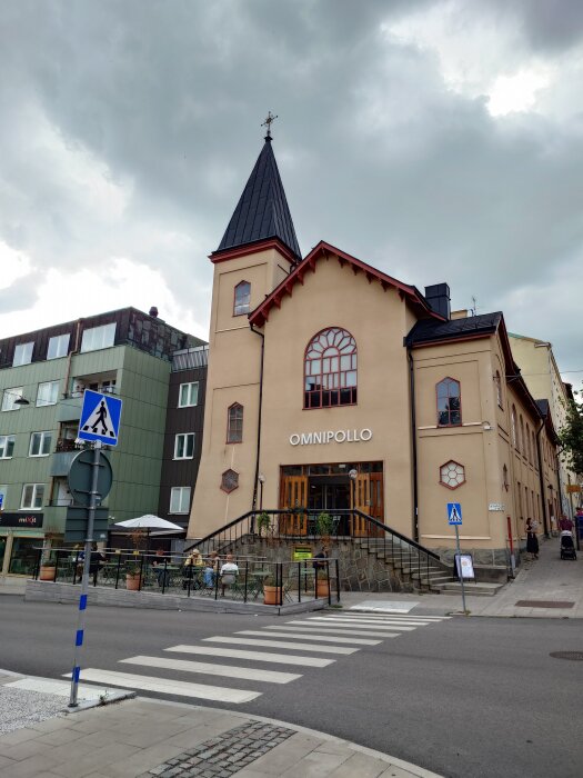 Gatuhörn med en byggnad märkt "Omnipollo", trottoar, övergångsställe och några uteserverade bord med människor under en molnig himmel.