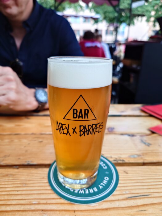 Ett glas öl märkt "Apex x Barrels" på ett träbord, med en person i bakgrunden.