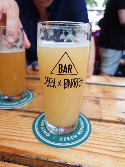 Ett glas ljus öl med logotypen "Apex x Barrels" på ett träbord, med en hand i bakgrunden som håller ett annat glas.