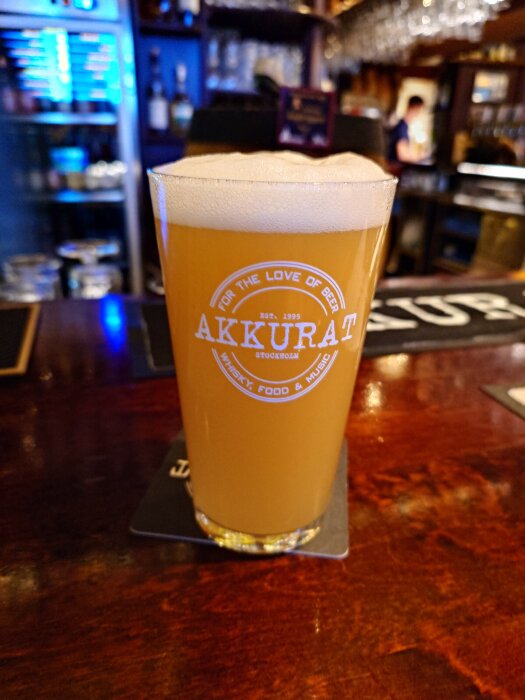 Ett glas öl med skumkrona på en bardisk, glaset märkt "Akkurat", i en bar med suddig bakgrund.