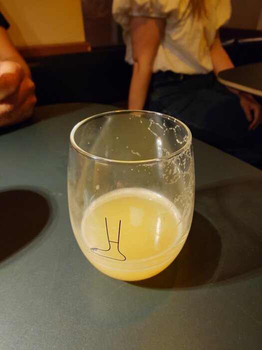 Ett nästan tomt glas med gul vätska, sannolikt en öl, på ett bord. En person sitter i bakgrunden.