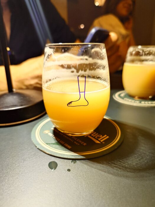 Ett glas med oklar guldfärgad dryck på ett bord, glaset har en illustration av en fot och står på en ölunderlägg.