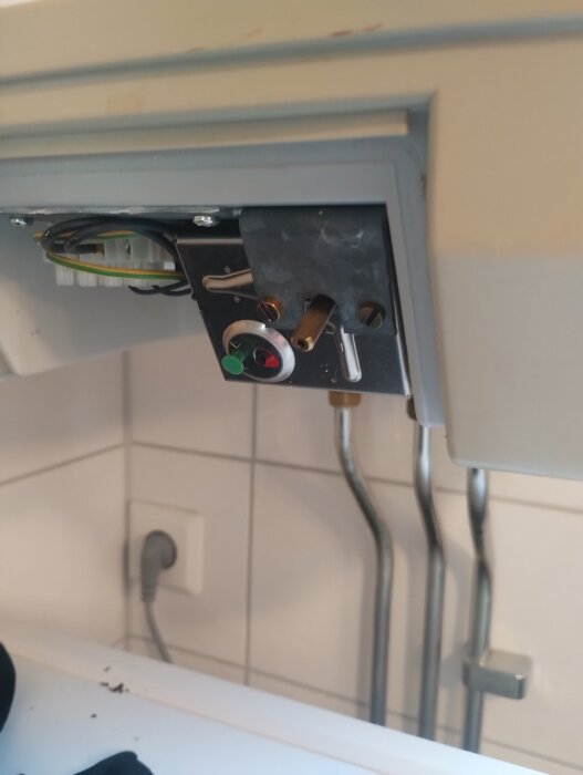 Närbild på insidan av en varmvattenberedare med en grön knapp, kablar och rör. Synliga vägguttag och rör i bakgrunden.