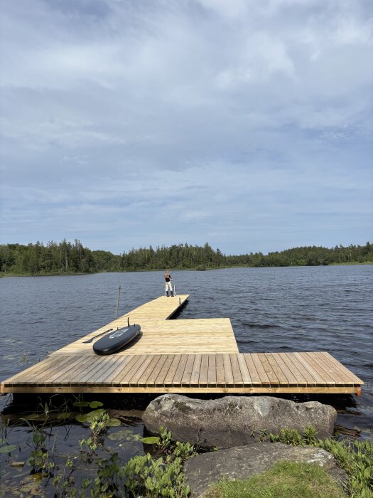 Nyrenoverad flytbrygga på sjö med stående person och en uppblåsbar båt på bryggan, omgiven av vatten och natur.
