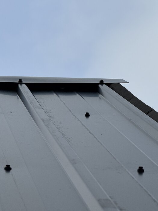 Böjd nockplåt monterad på brant plåttak, där takplåten inte når nock helt, med 10 cm papp under nockplåten som överlappar något över plåten.