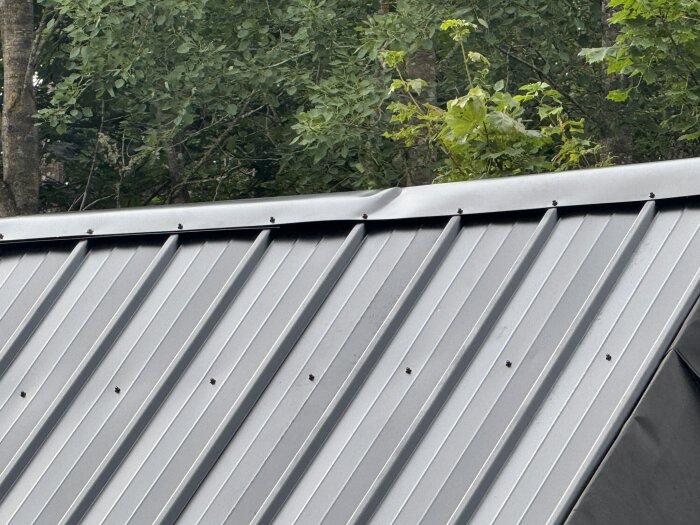 Nockplåt monterad på ett plåttak med synliga skruvar och böjda kanter. Grönska i bakgrunden och ingen nockplåtsstöd synligt under plåten.