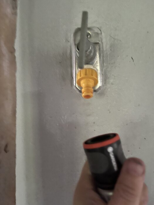 En hand håller i en svart och röd slangkoppling mot ett gult väggfäste, sannolikt orsakar vattenläckage.