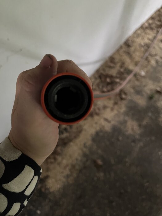 Hand håller i en orange trädgårdsslangkoppling. Bakgrunden är oskarp och visar mark och vägg.