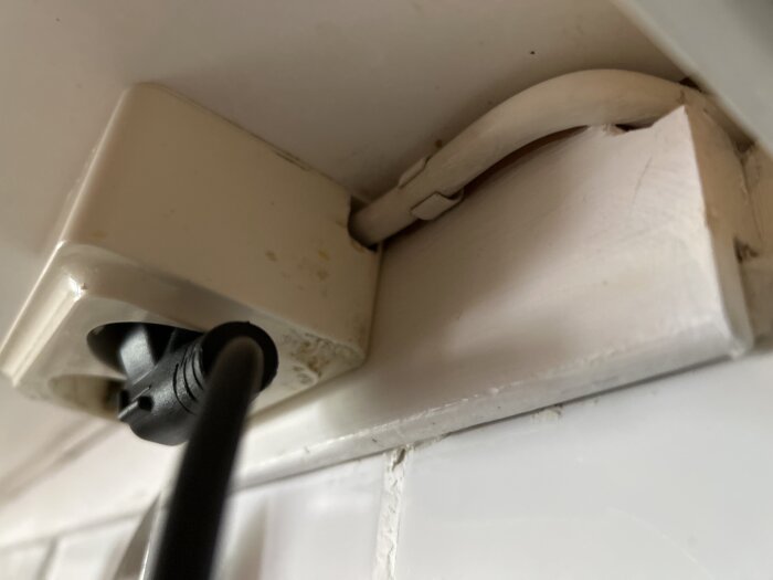 Vägguttag ovanför spishäll med borrat hål för kabel, nära köksfläkt. Svart sladd ansluten i uttaget, synlig vit kabel till lampa.