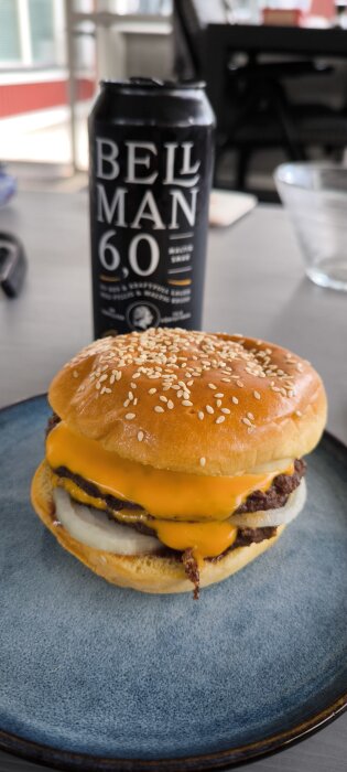Hamburgare med smält ost och lök i ett sesambröd på en tallrik, med en burk öl i bakgrunden.