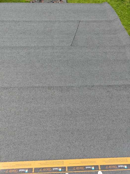 Grått tak med flera lager av takpapp och tydliga överlappningar, del av ett byggprojekt i trädgårdsmiljö.
