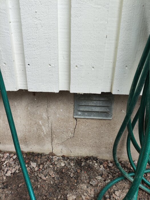 Spricka i betonggrund under enplanshus med vit träpanel och ventilationsgaller. Grön vattenslang synlig framför sprickan.