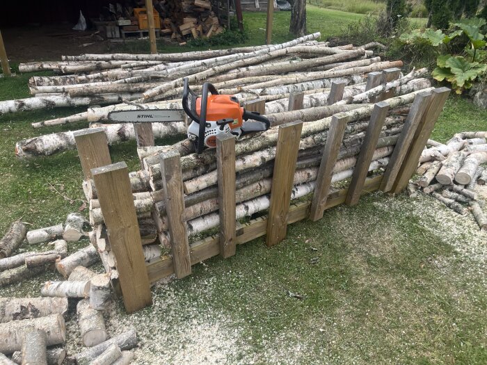 Hyvelbänk med staplade vedträn och en motorsåg ovanpå, omgiven av fler vedträn på gräs.