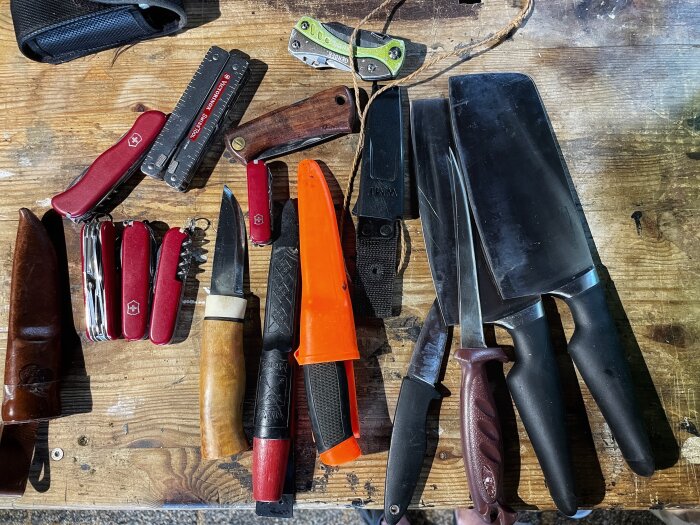 Olika typer av knivar och multiverktyg ligger utspridda på ett träbord, inklusive flera röda schweiziska arméknivar och stora kockknivar med svarta handtag.