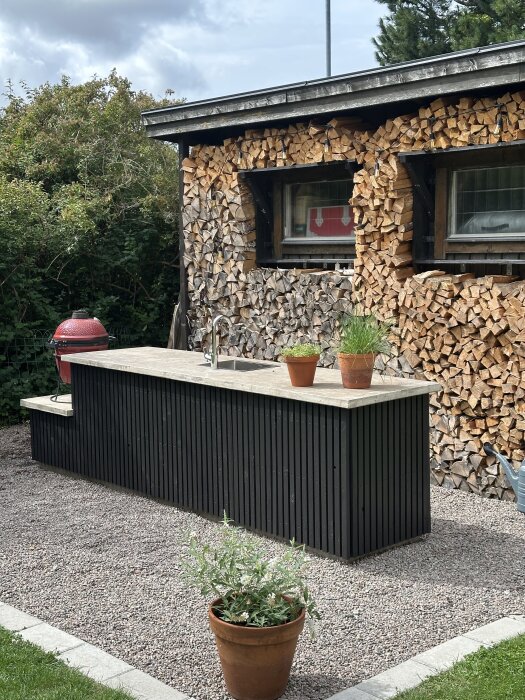Utekök med svart ribbad front och betongbänkskiva, inklusiv diskho och kran. Bilden visar även en stapel ved, växter i krukor och en grill.