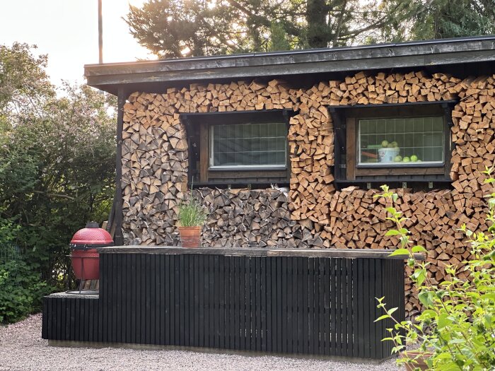 Svartmålat utekök med ribbor framför staplat ved, röd grill och krukväxt ovanpå. Träd och buskar i bakgrunden.