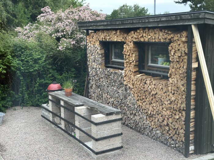 Utekök med lecablock, träklädd vägg med vedförvaring och blommande buskar. Grill och blomkruka på arbetsbänken.