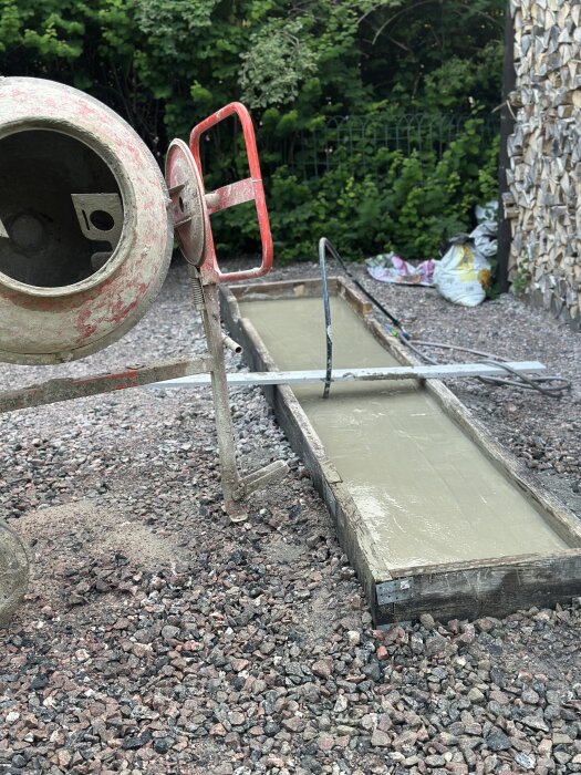 Betongblandare och nylagd betongplatta i en form. Omgiven av grus och stenar, med träd och buskar i bakgrunden.