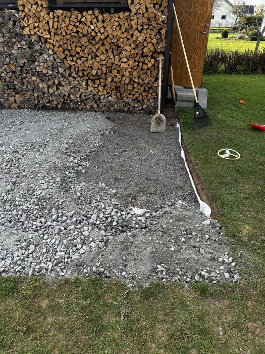 Grus och bergskross på markduk vid bygge av utekök. Ved staplad mot väggen, spade och kratta lutade mot huset, gräsmatta i bakgrunden.