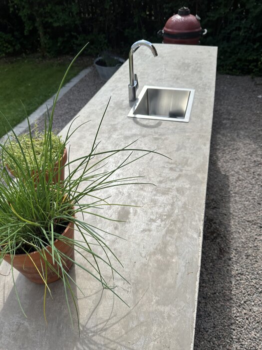 Betongbänkskiva med inbyggd diskho och blandare i ett utekök, omgiven av en kruka med gräslök och en röd grill i bakgrunden.