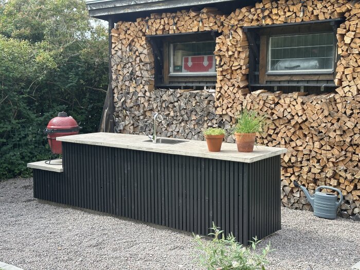 Svart ribbat utekök med betongbänkskiva och diskho, omgiven av staplad ved. Två krukväxter och en röd grill syns på sidan.