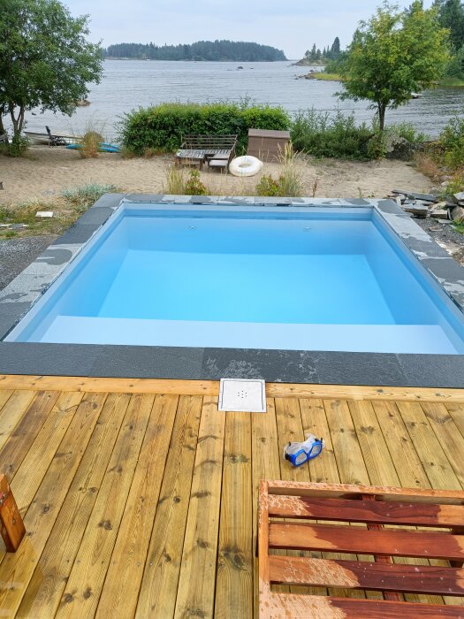 Träaltan med nybrädor i nivå med poolkant, placerad vid vatten med utsikt över sjö och träd i bakgrunden. Simglasögon ligger på trallen.