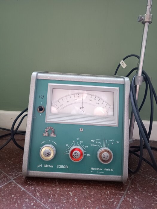 Gammal pH-mätare med analoga mätare och knappar, märkt "Metrohm E350B", med kablar mot en grön vägg.