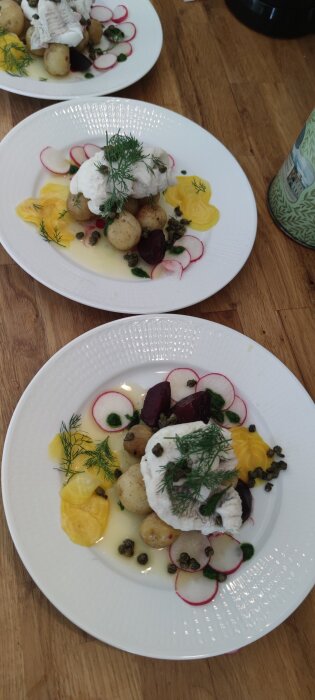 Två tallrikar med kolja, potatis, rädisor, gul rödbeta, kapris och dill serverade på ett träbord.