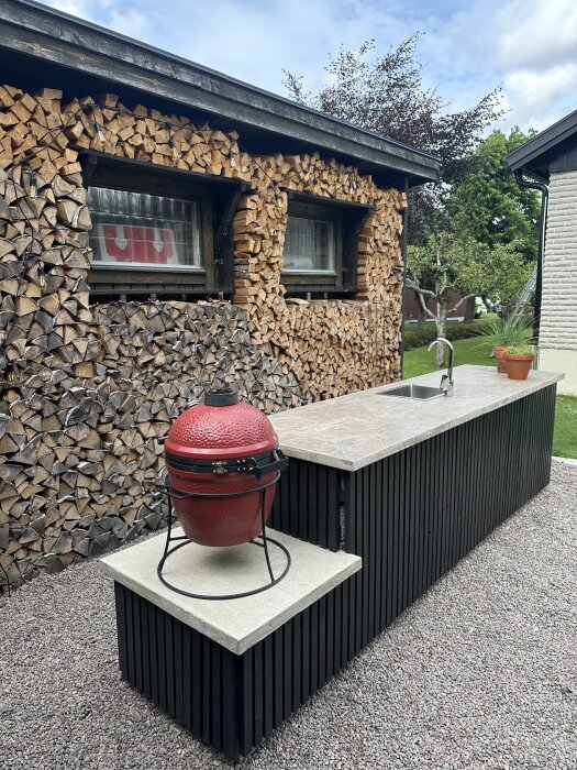 Utomhuskök med röd kamado-grill, handfat, vedförråd och terrass med betong- och träytor, omgiven av gröna växter och grus.