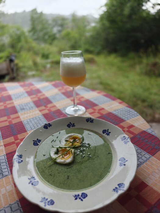 Nässelsoppa med kokt ägg dekorerad med gräslök serverad på en dekorativ tallrik, med ett glas dryck i bakgrunden på en rutmönstrad duk.