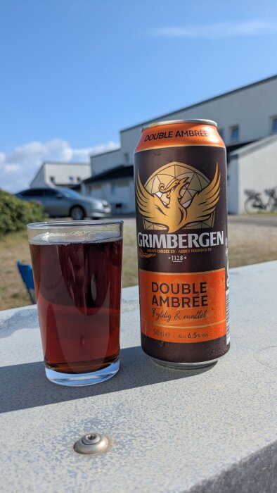 En burk Grimbergen Double Ambree och ett glas med samma dryck står på ett bord utomhus, med ett hus och blå himmel i bakgrunden.