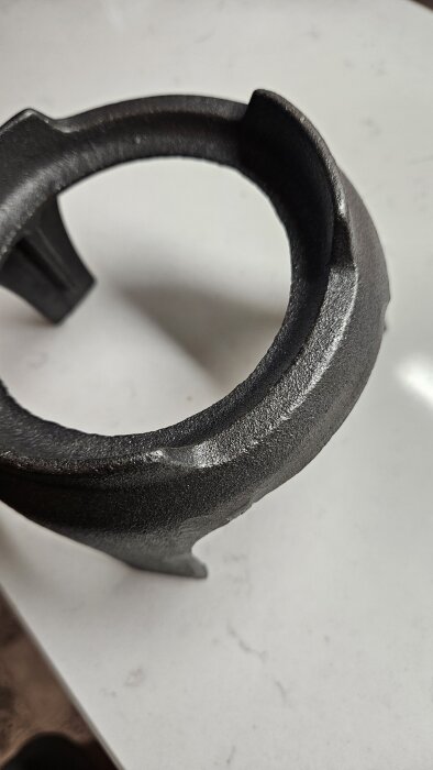 Svart metallring, eventuell del av en gryta eller ställning, på ljus yta. Diskuteras för justering med vinkelslip och möjligt rostskydd.