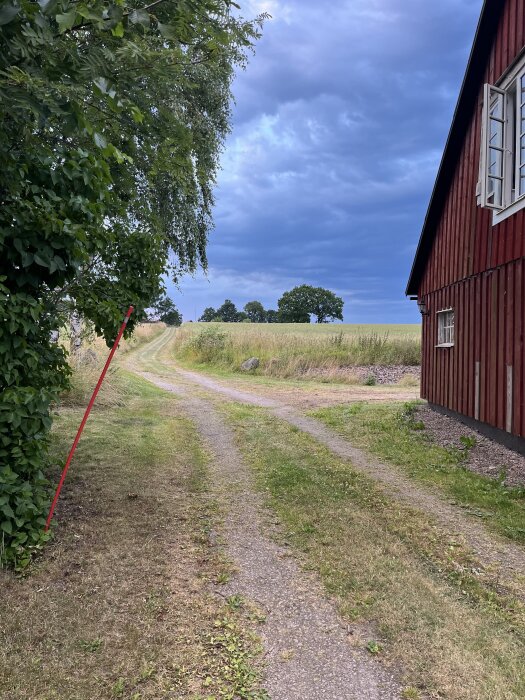 Landsväg vid röd träbyggnad, grönt fält och kraftigt molntäcke.