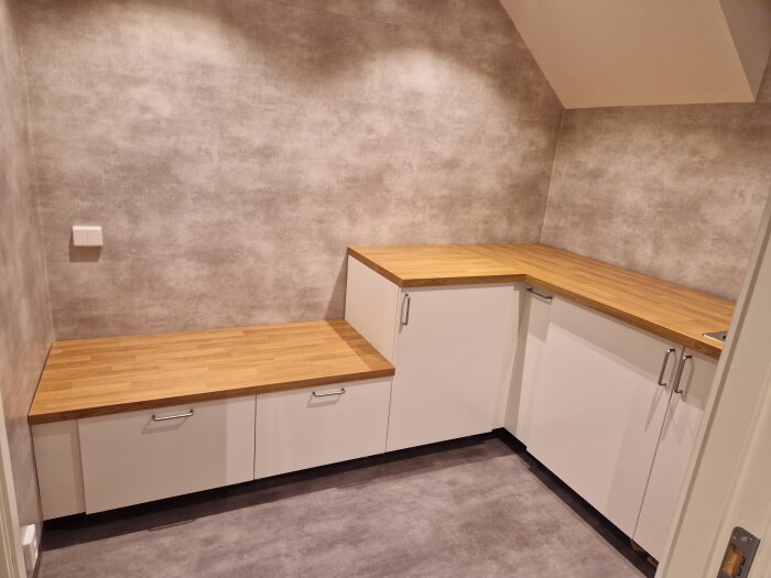 Tvättstuga under renovering med träbänkar och vita skåp i hörnet, väntar på rörmokare för slutförande.